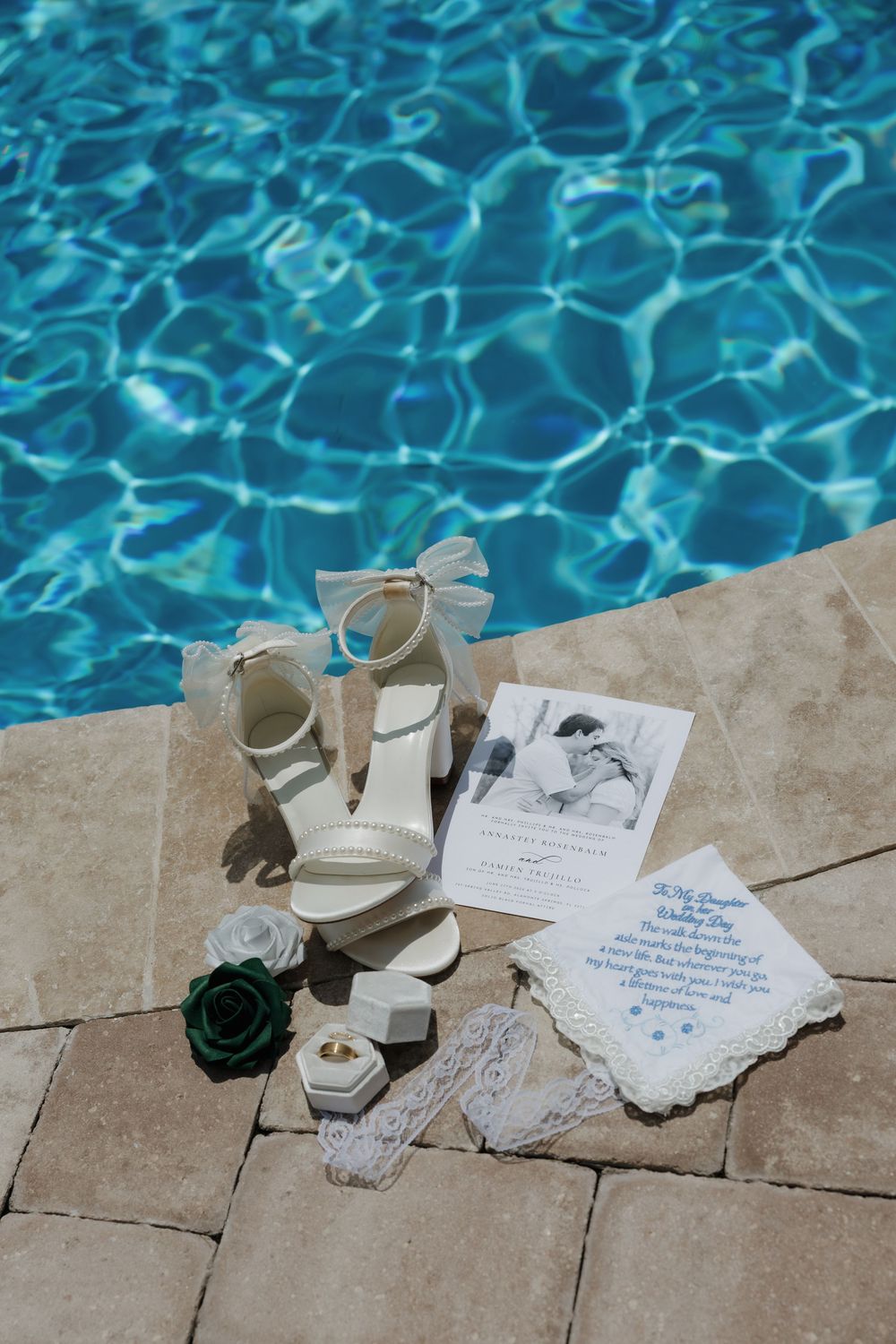 Bridal flat lay featuring white heels, wedding stationery, lace handkerchief, rings, and green roses by a sparkling blue pool.