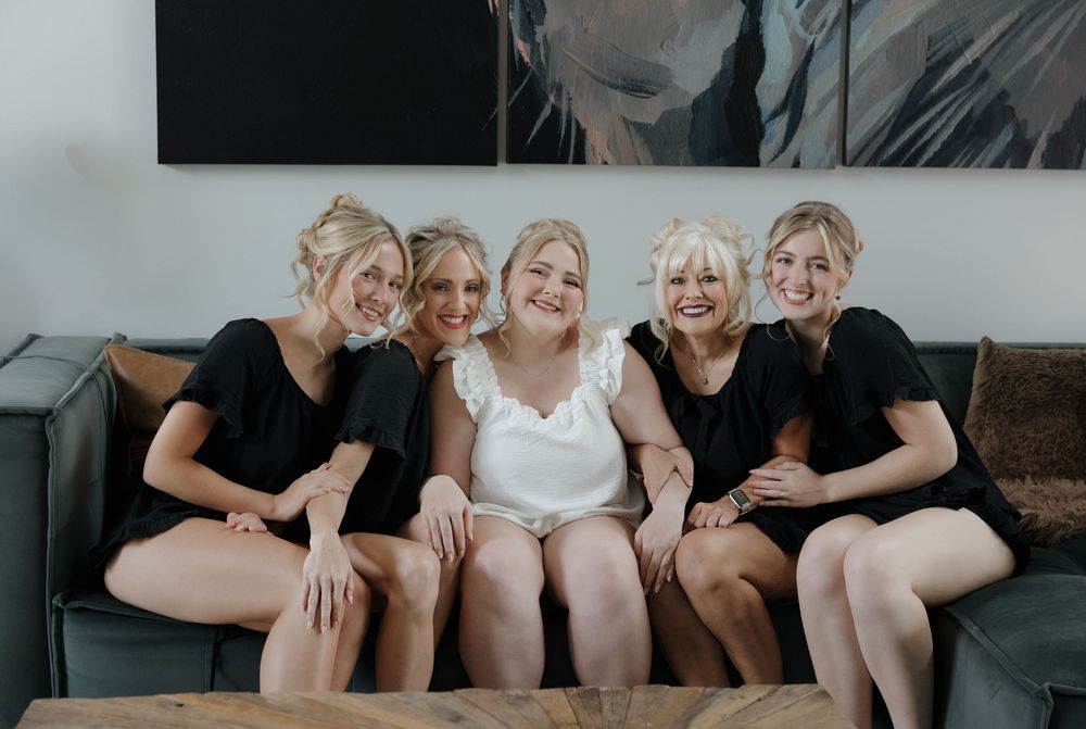 Bride in white with four bridesmaids in black dresses sitting together on a grey sofa, smiling and laughing.
