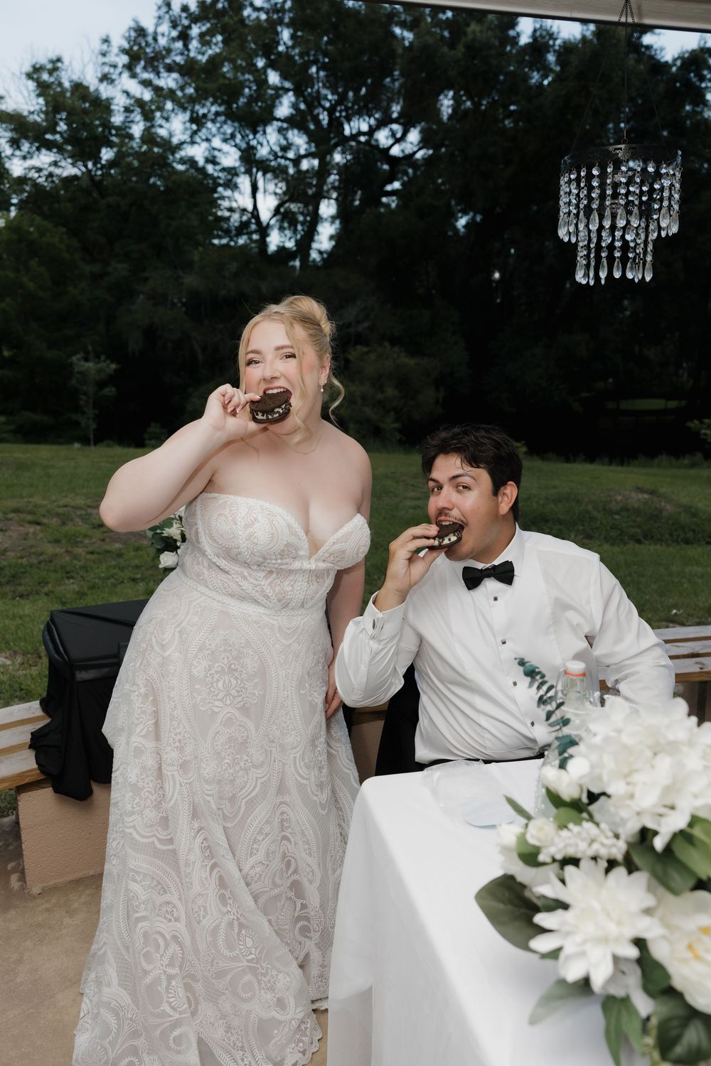 Bride and groom eating chocolate donuts together at outdoor wedding reception, bride in lace gown, groom in tuxedo.