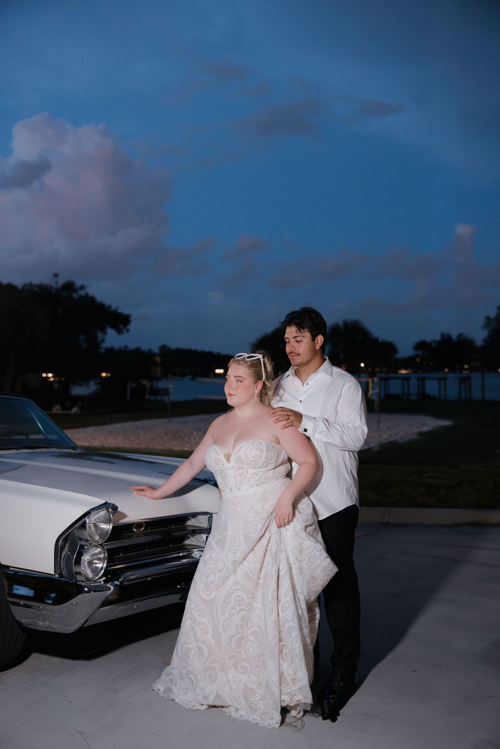 Bride and groom pose beside a classic white car at dusk, bride in lace gown, groom in white shirt.