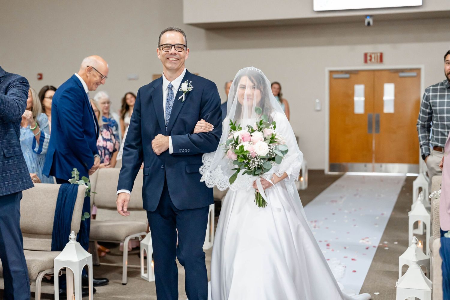 Bride in white gown and veil walks down the aisle with escort holding bouquet at elegant church wedding ceremony.