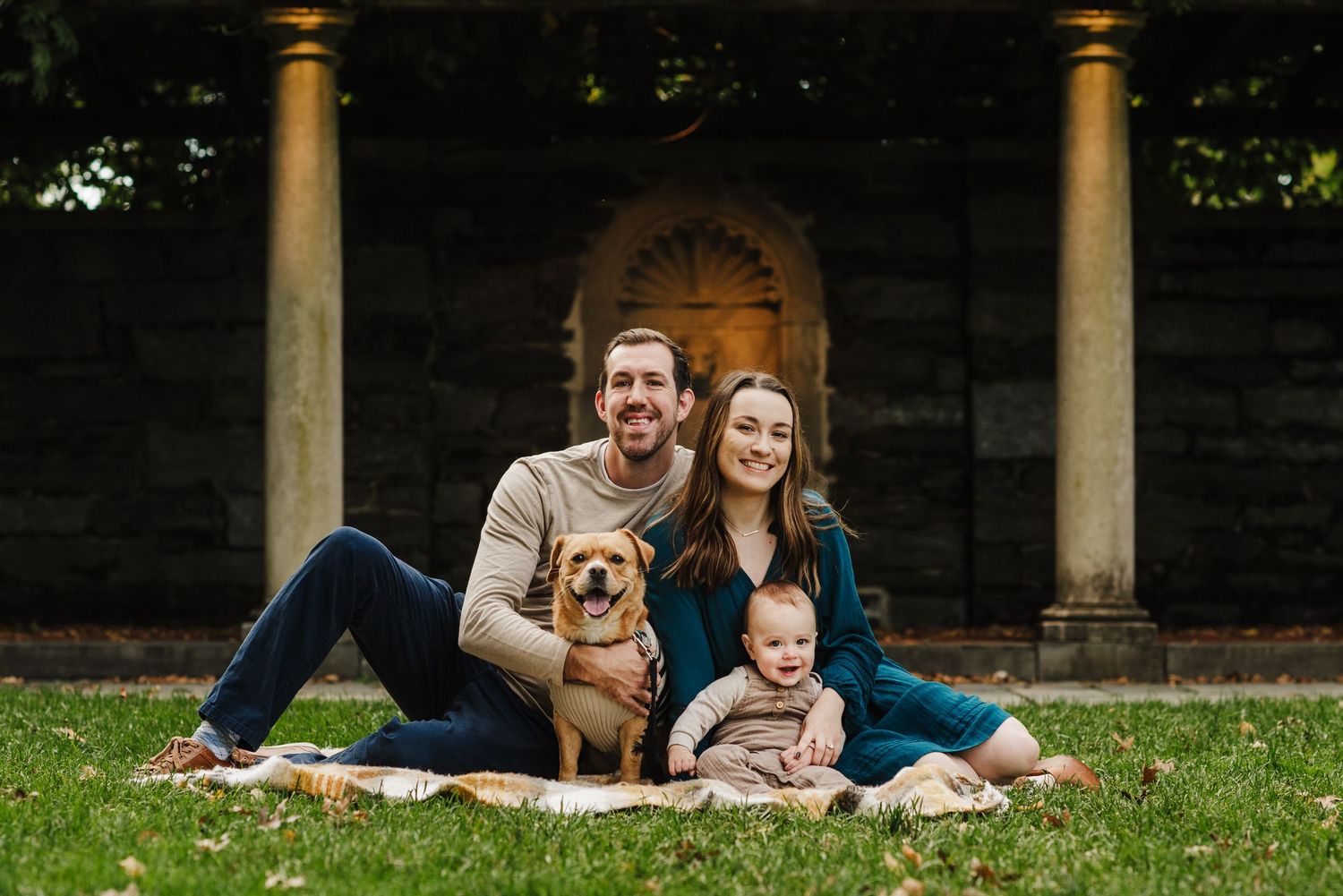 A smiling couple sits on a blanket outdoors with a golden retriever and baby in front of stone columns.