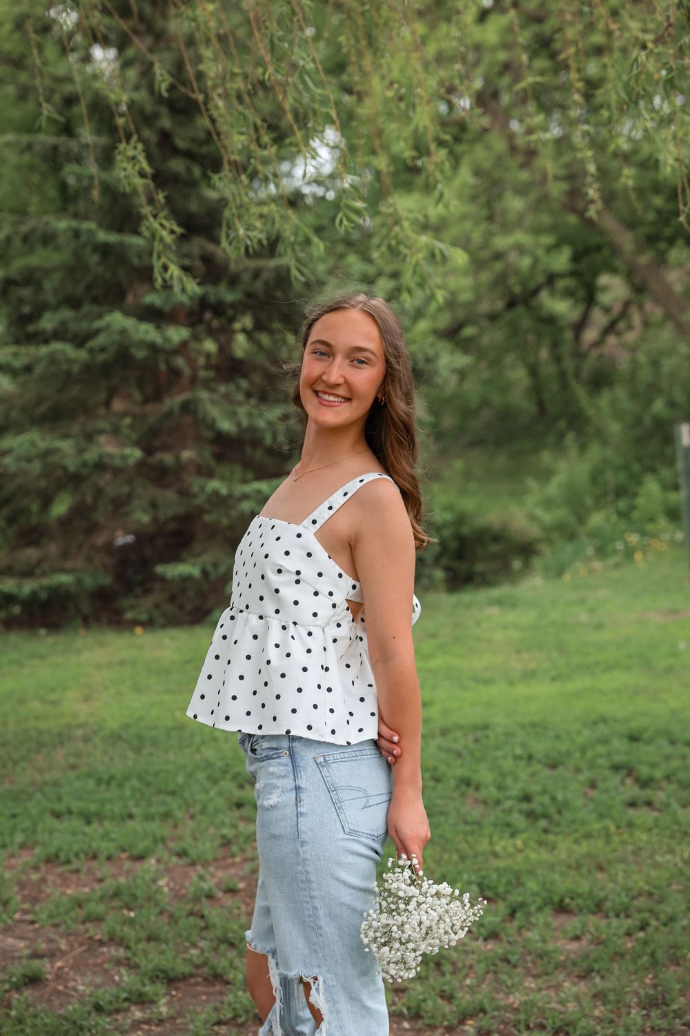 Smiling teenage girl in polka dot top and jeans holding white flowers outdoors.