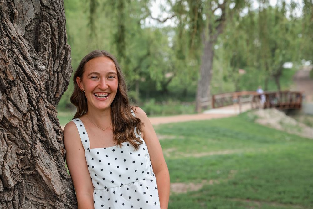 Young woman smiling beside a tree in a lush green park, wearing a white polka dot dress on a sunny day.