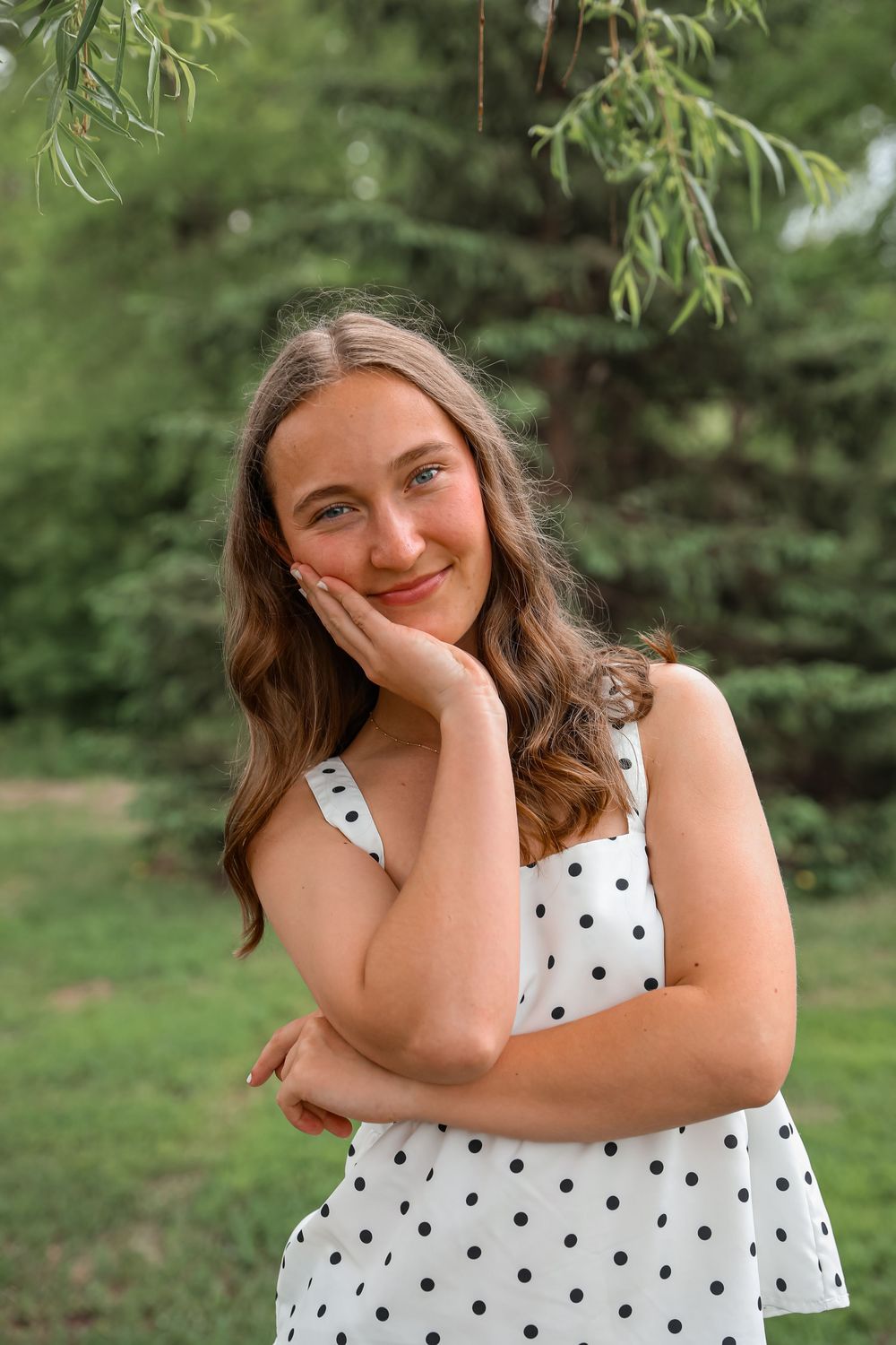 Smiling teenage girl in white polka dot dress posing outdoors with green trees in background.