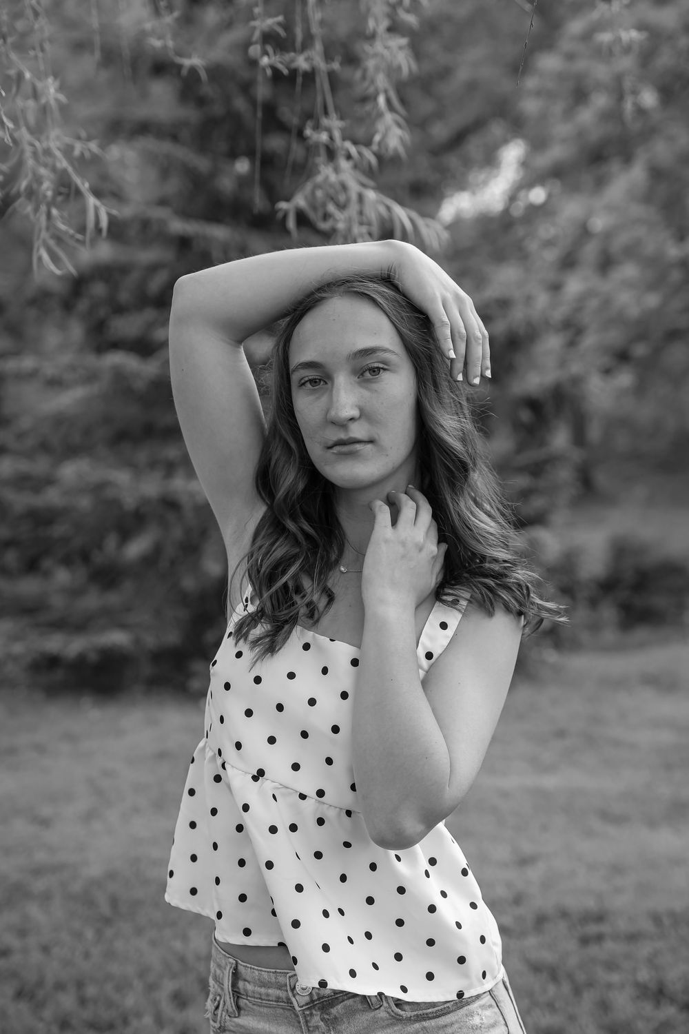 Black and white portrait of a young woman in polka dot top posing outdoors with trees in background.