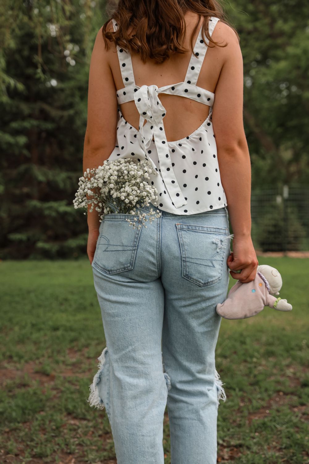 Young woman in polka dot top and jeans holding flowers and stuffed animal in park setting.