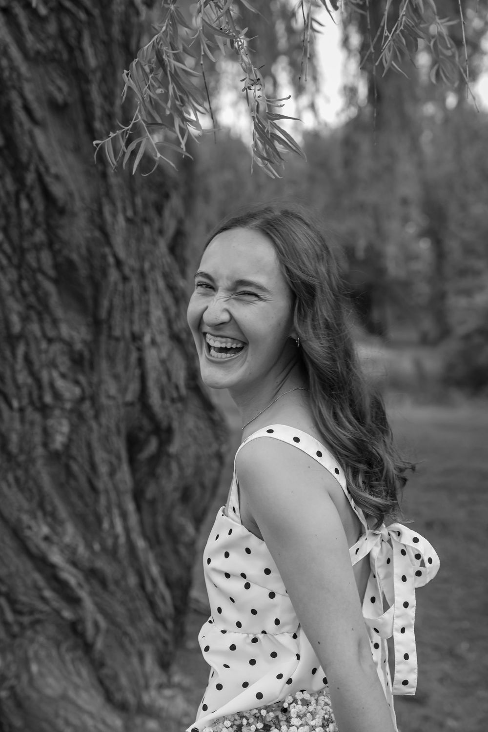 Black and white portrait of a laughing young woman in polka dot dress leaning against a tree outdoors.