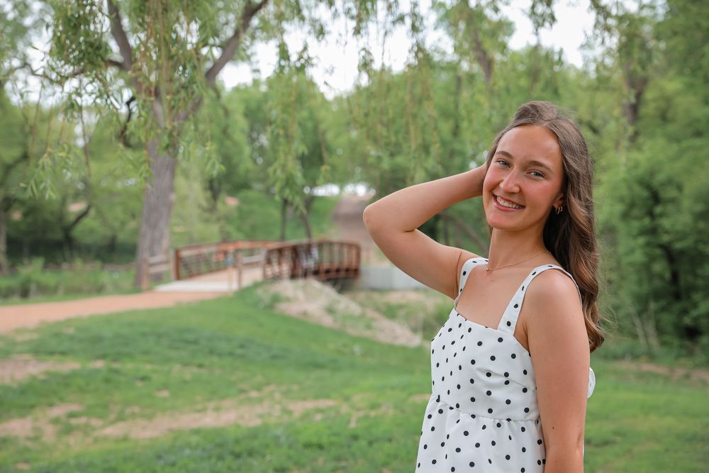 Young woman in polka dot dress smiling outdoors in a lush green park with a wooden bridge.