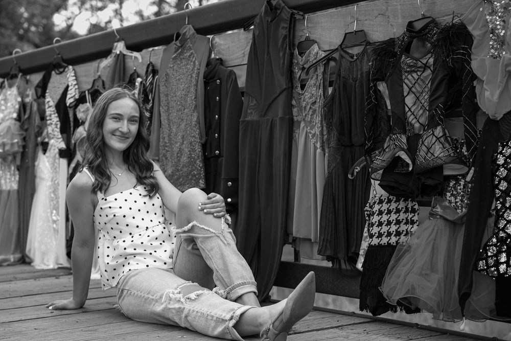 Young woman sitting on wooden deck beside hanging dresses at outdoor clothing display, black and white photo.