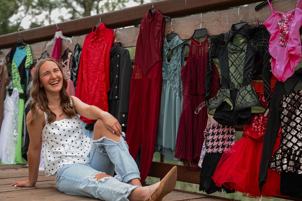 A smiling woman sits on a deck surrounded by colorful dance costumes hanging on a rack outdoors.