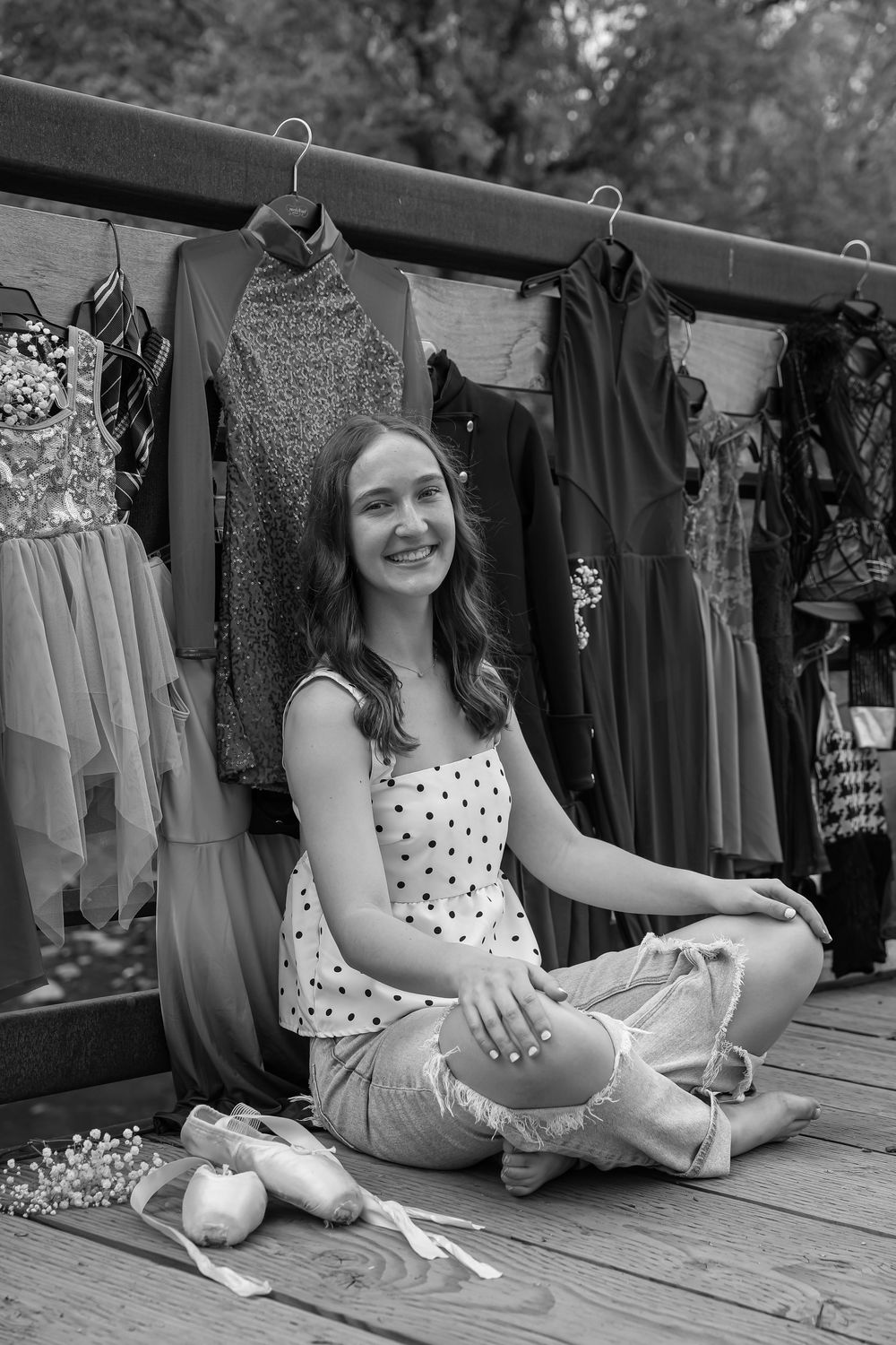 Black and white photo of a smiling young woman sitting cross-legged on a deck surrounded by hanging dresses and ballet shoes.