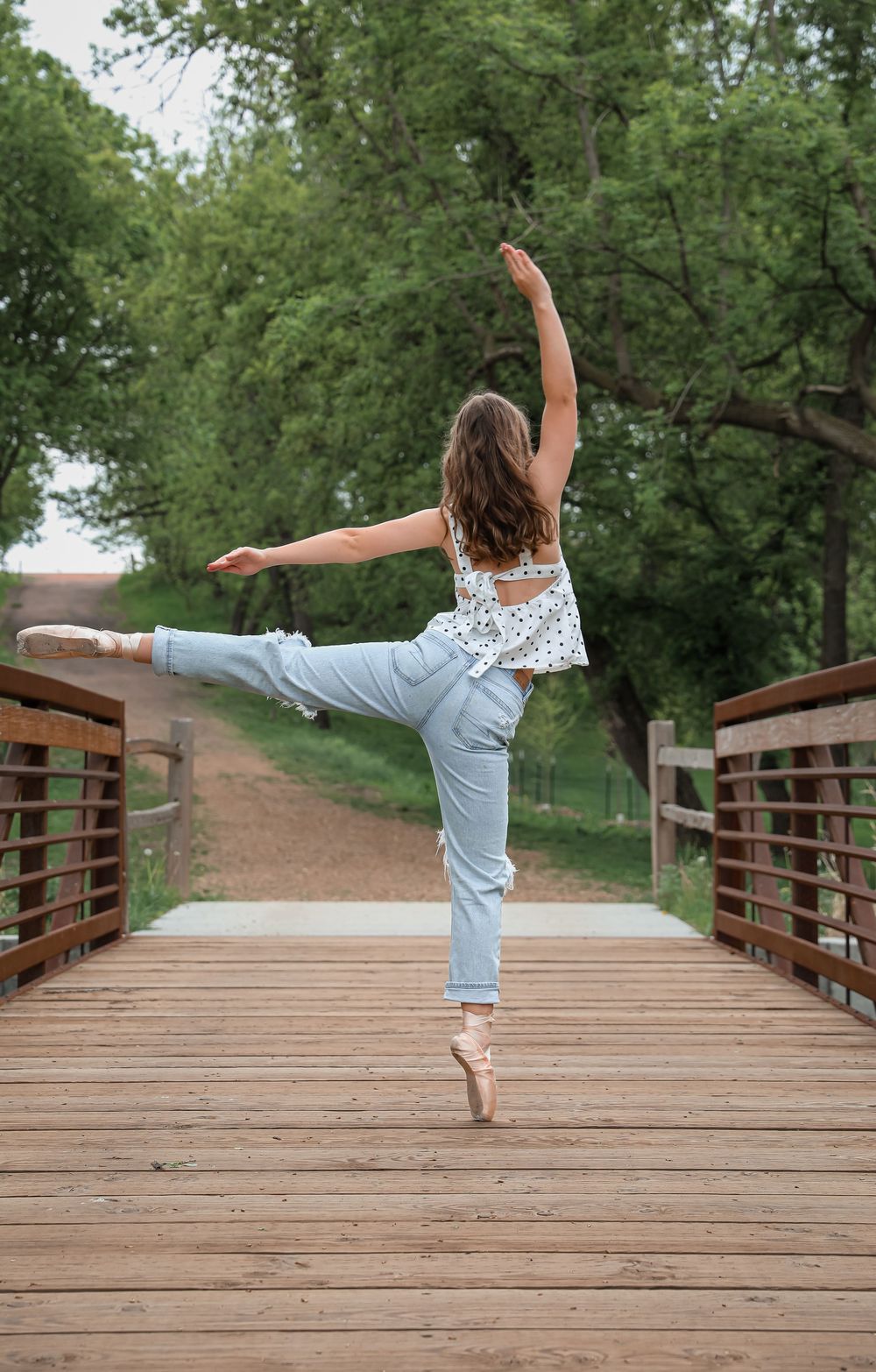A ballet dancer poses gracefully in pointe shoes on a wooden bridge surrounded by green trees.