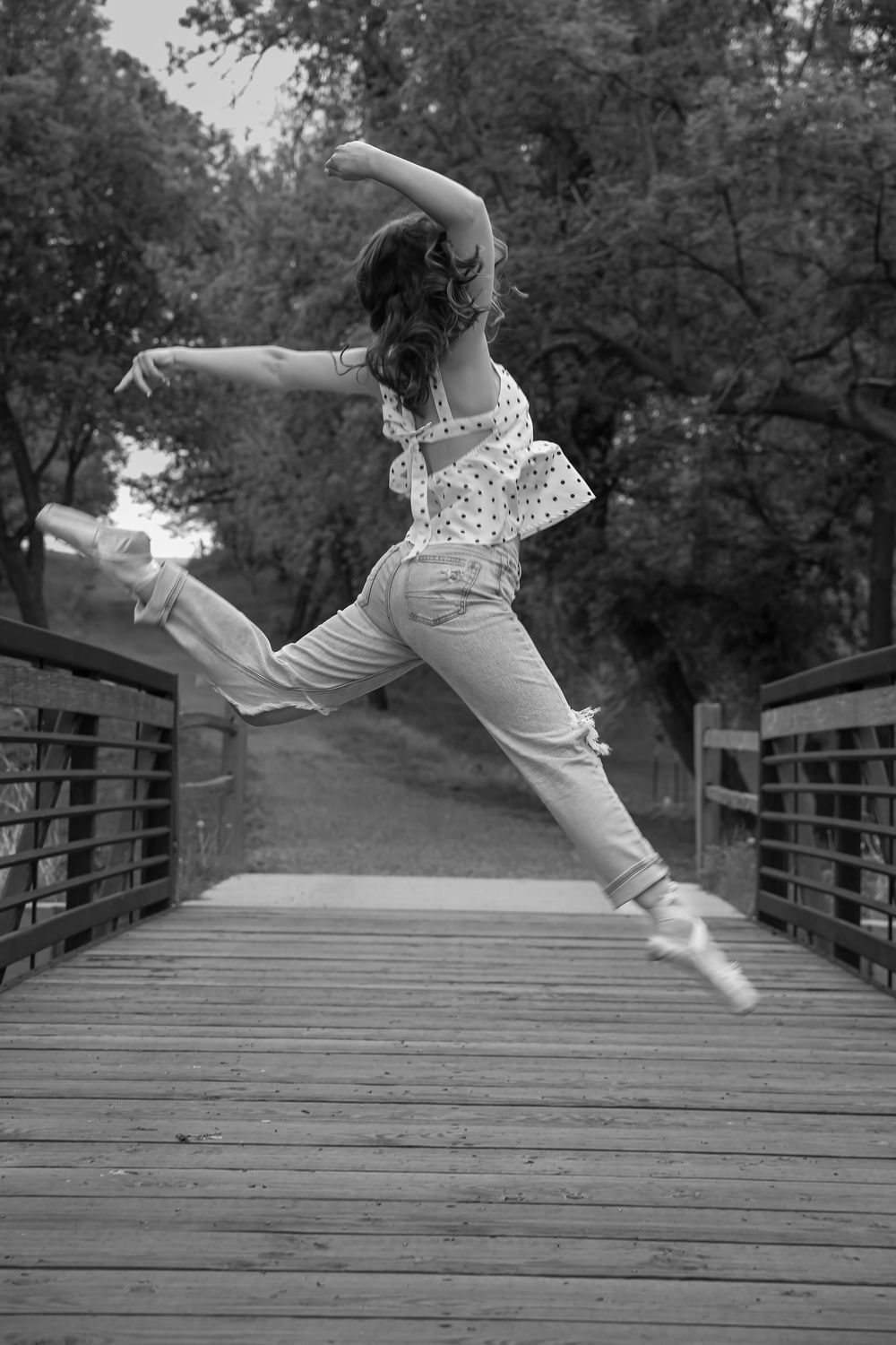 Black and white photo of a ballet dancer leaping gracefully on a wooden bridge wearing jeans and pointe shoes.