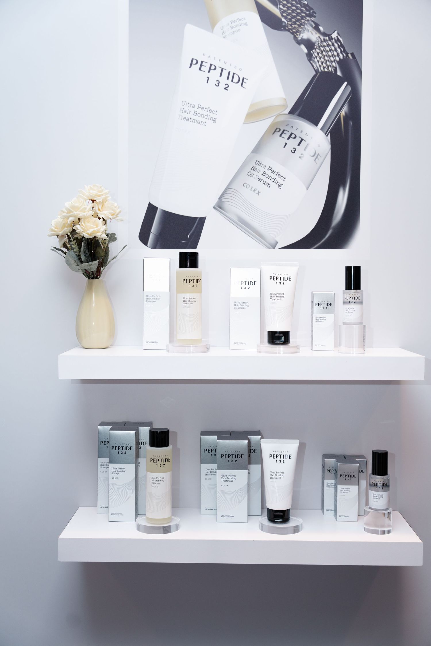 White shelves displaying Peptide 132 skincare products with a promotional poster, beside a vase of white flowers.