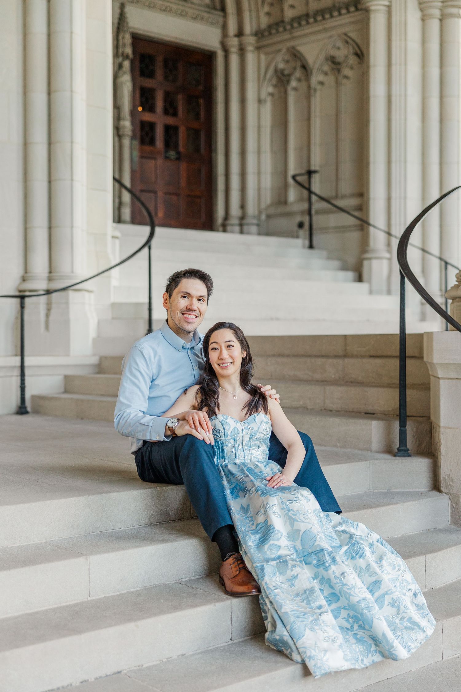 Couple sitting on stone steps of elegant building, woman in blue floral dress, man in light blue shirt, smiling.