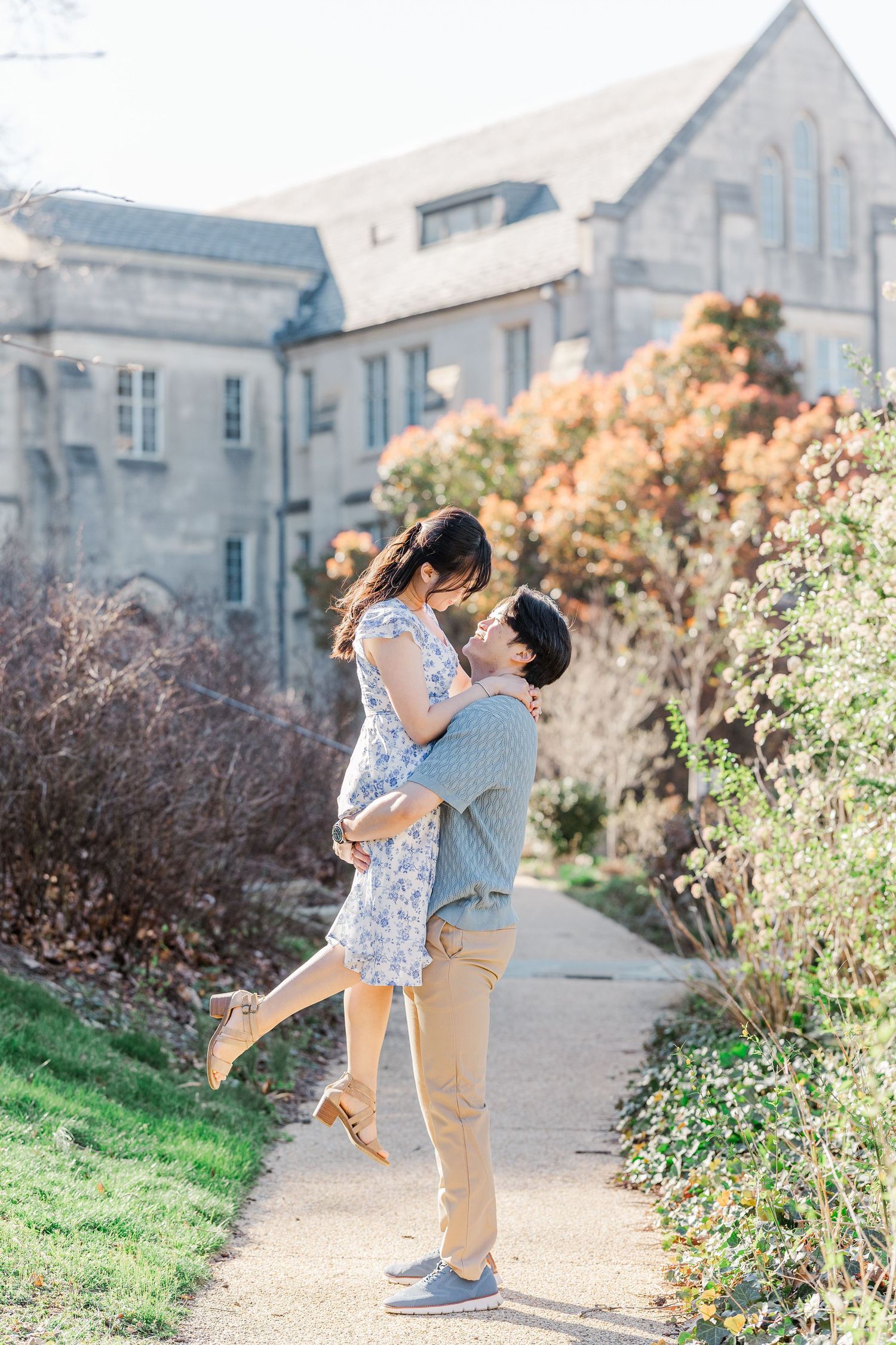 A man lifts a woman for a kiss on a garden path with a stone building and autumn shrubs in the background.