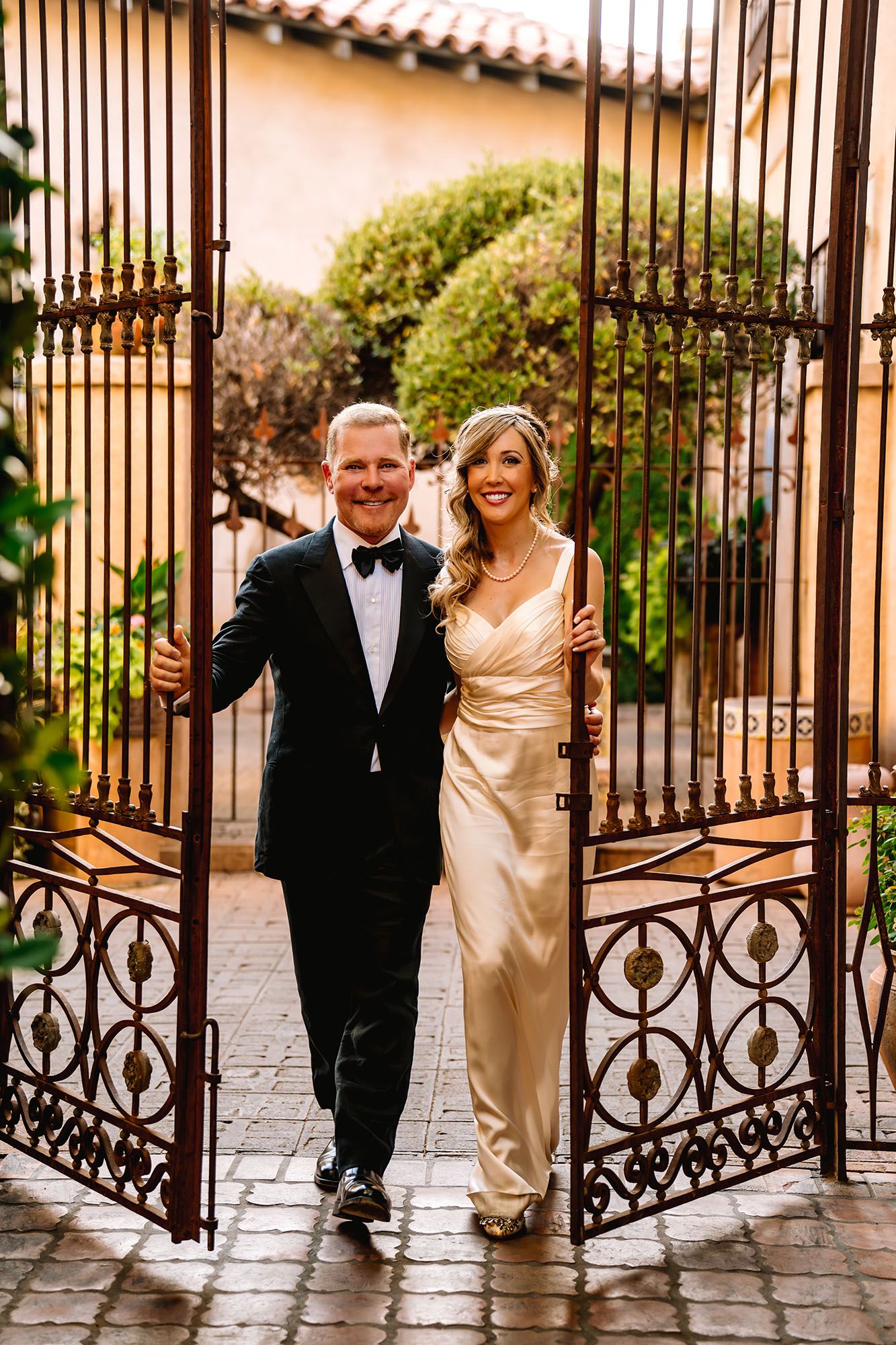 Smiling couple in formal attire walking through ornate iron gates at an elegant outdoor venue.