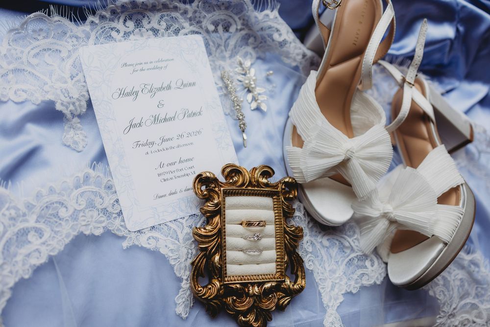 Wedding flat lay with white heels, gold framed rings, floral invitation, and blue lace fabric on elegant bridal display.