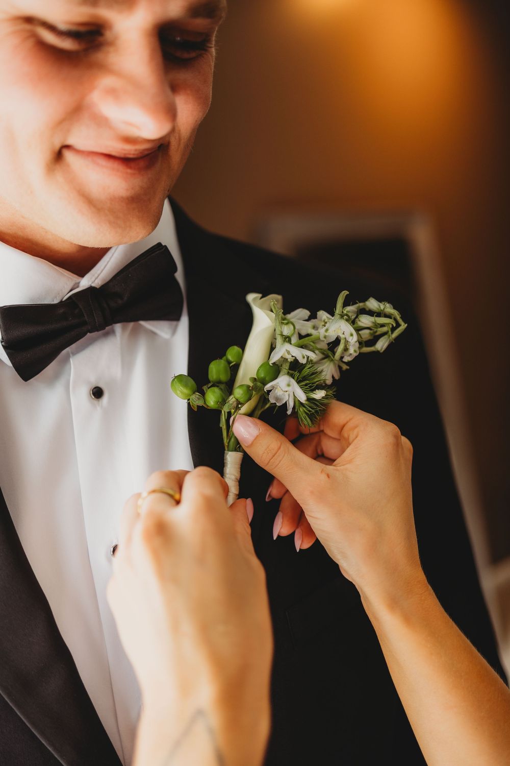 Hands pinning a white floral boutonniere on a smiling groom in a black tuxedo with bow tie.