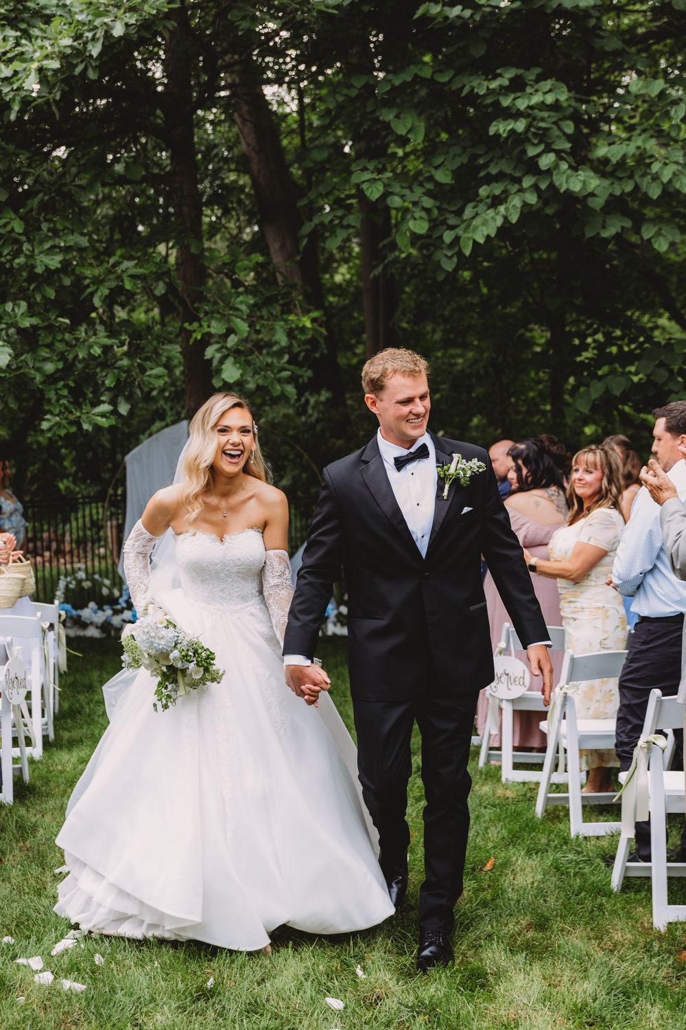 Joyful bride and groom walk down outdoor wedding aisle, bride in strapless lace gown, groom in black tuxedo.