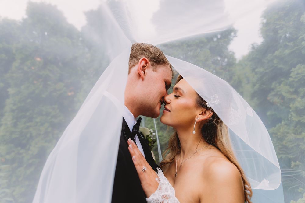 A bride and groom share a romantic kiss under a flowing white veil on their misty wedding day.