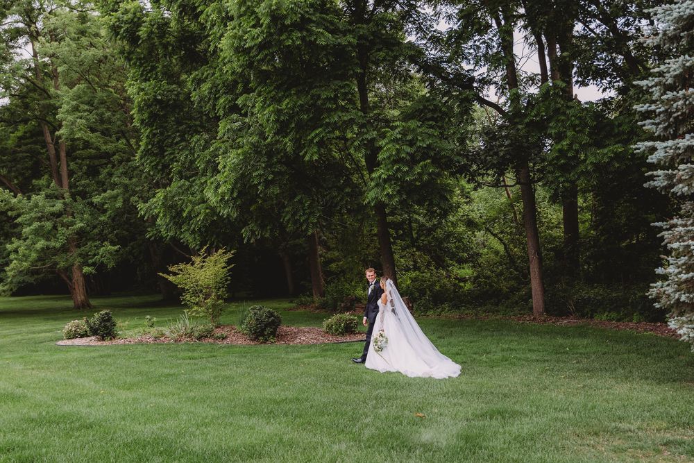 A bride and groom share a romantic moment on a lush green lawn surrounded by tall trees at an outdoor wedding.