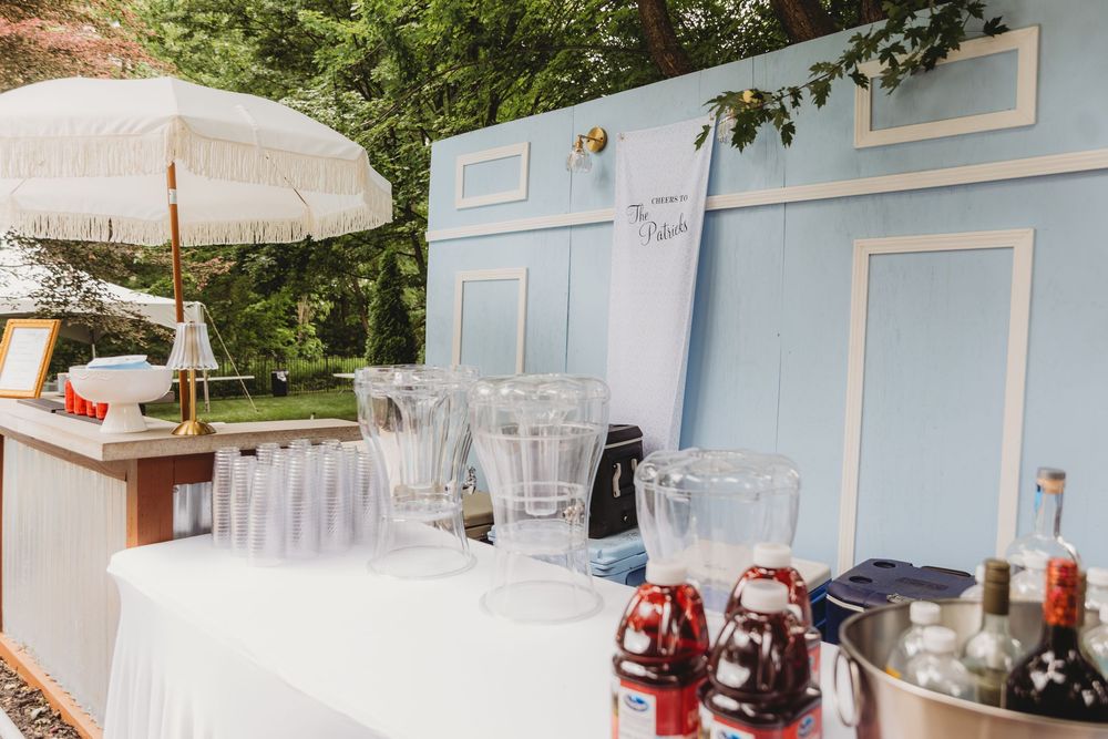 Outdoor bar setup with glassware, pitchers, and colorful drink dispensers on a white-draped table beside a light blue backdrop.