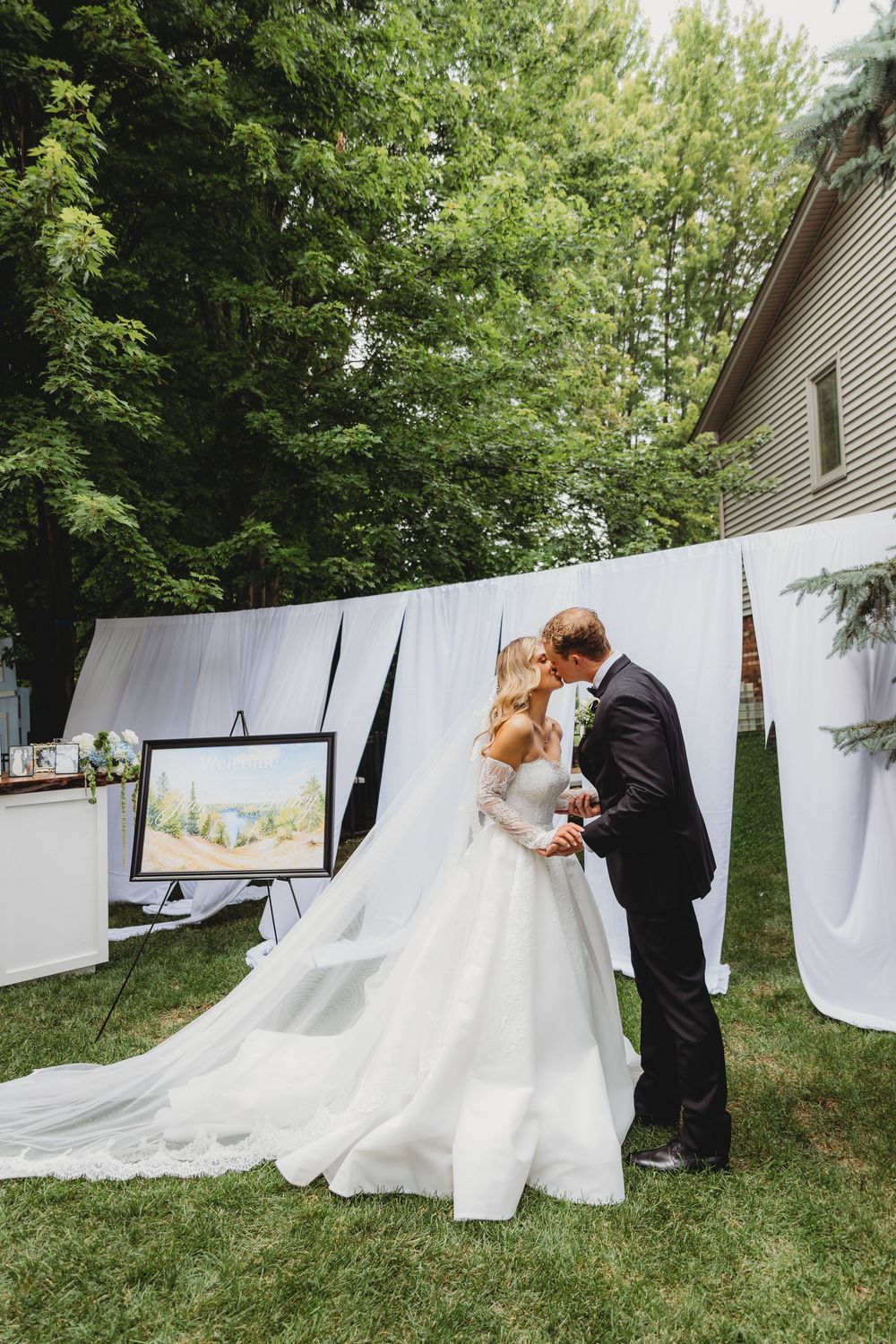 Bride and groom share a kiss outdoors at a backyard wedding ceremony with white draping and lush green trees.