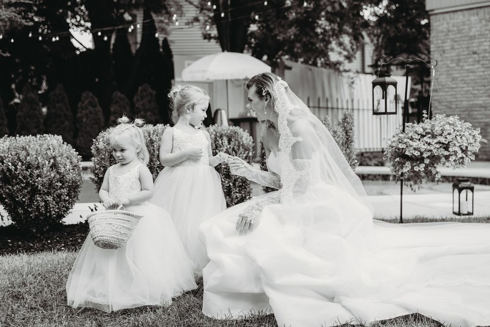 Black and white photo of a bride in a wedding gown kneeling with two young flower girls outdoors.