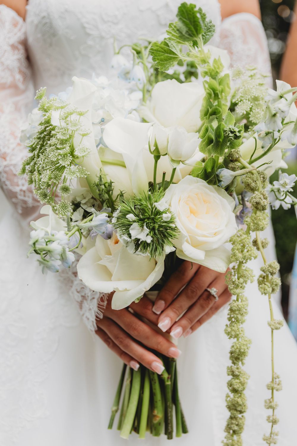 Bride holding a lush white and green wedding bouquet with roses, tulips, and wildflowers, wearing a lace gown.