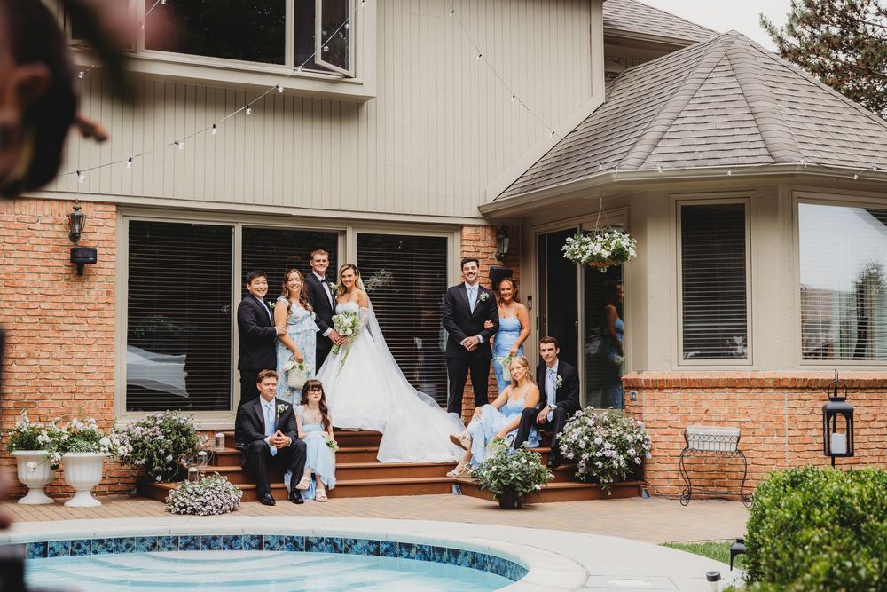 Wedding party poses outside brick home near pool, bride in white gown surrounded by family in blue attire.