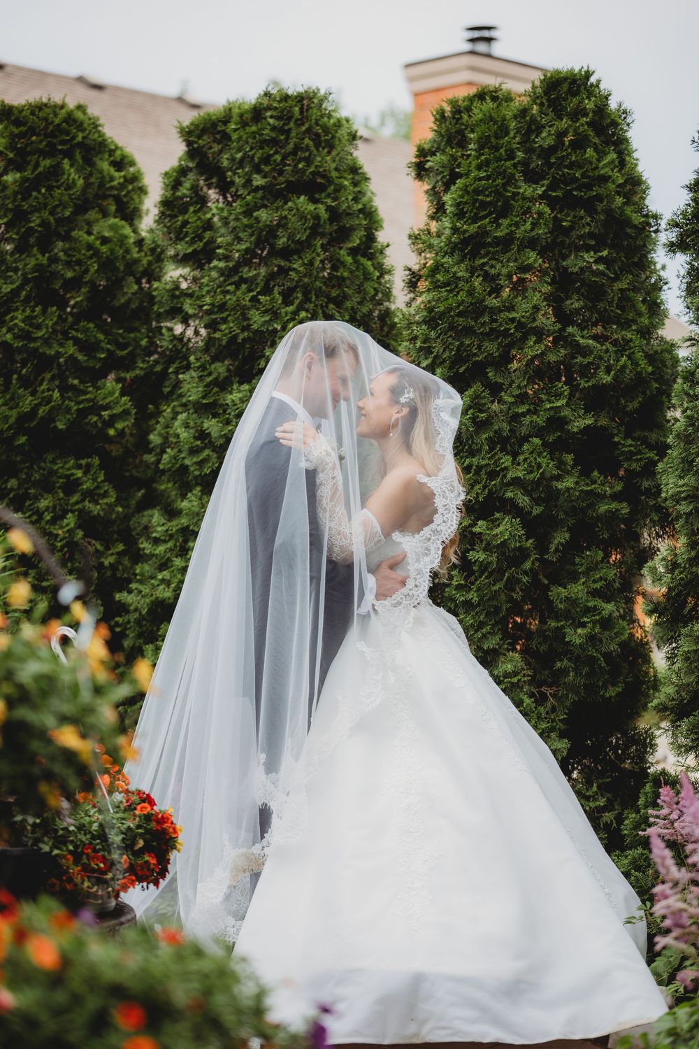 Bride and groom sharing a romantic kiss under a flowing white veil in a lush garden setting.