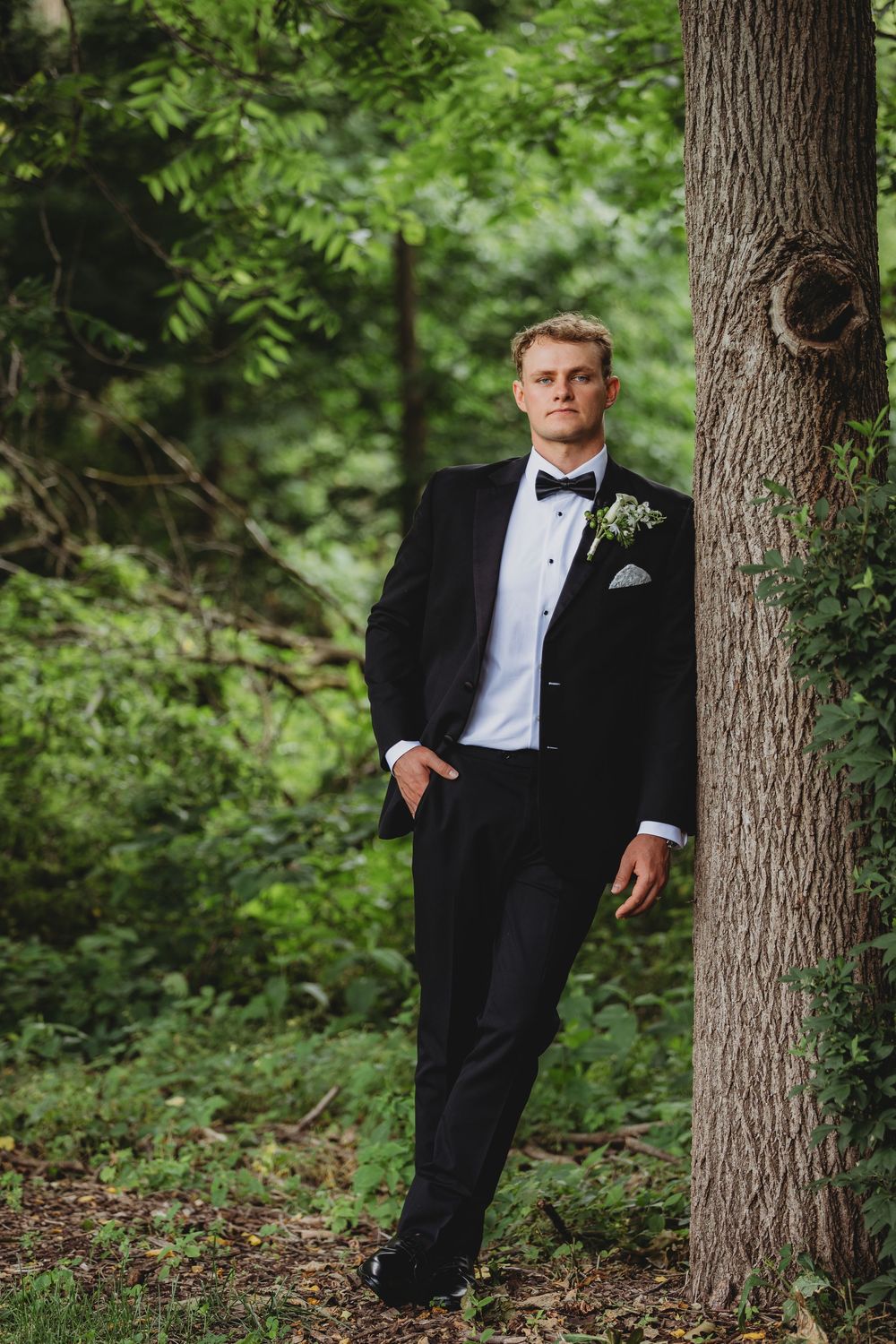 Groom in black tuxedo with boutonniere leans against tree in lush green forest setting.