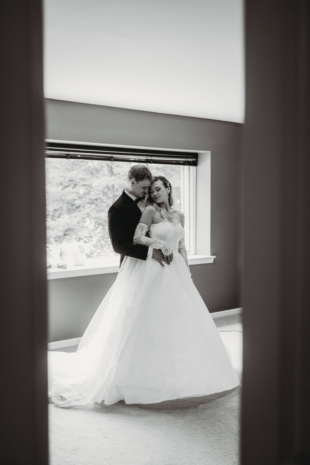 Black and white photo of bride and groom embracing tenderly by window, bride in elegant ballgown.