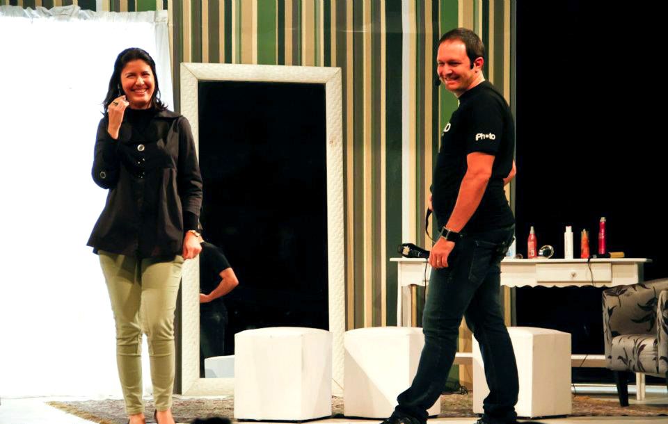 Two performers smile on a theater stage with striped walls and white furniture props.