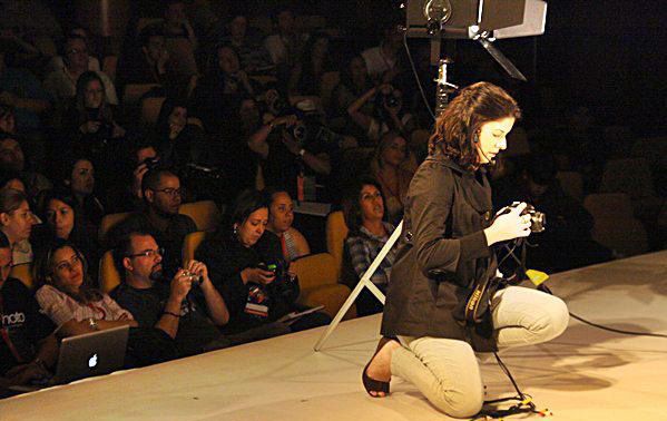 Musician kneels on stage playing guitar before a large audience at an indoor concert event.