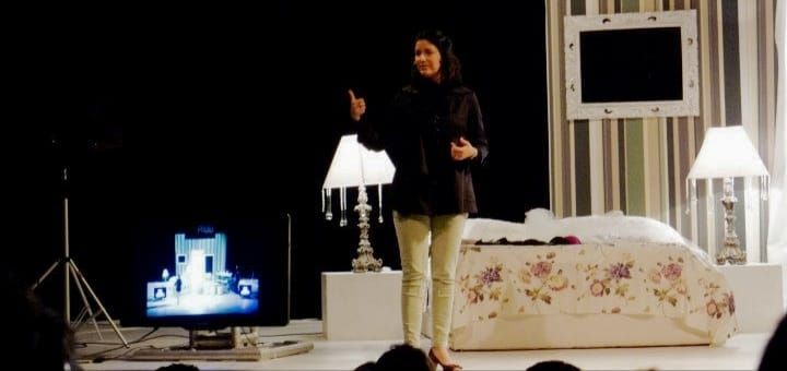 A woman speaks on stage near a floral bed, lamp, and TV in a theatrical bedroom set design.