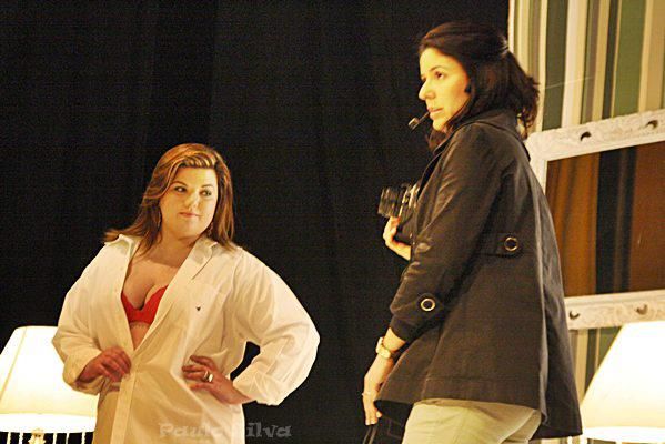 Two women stand on a stage during a theatrical rehearsal, one in a white coat and one in a dark jacket.