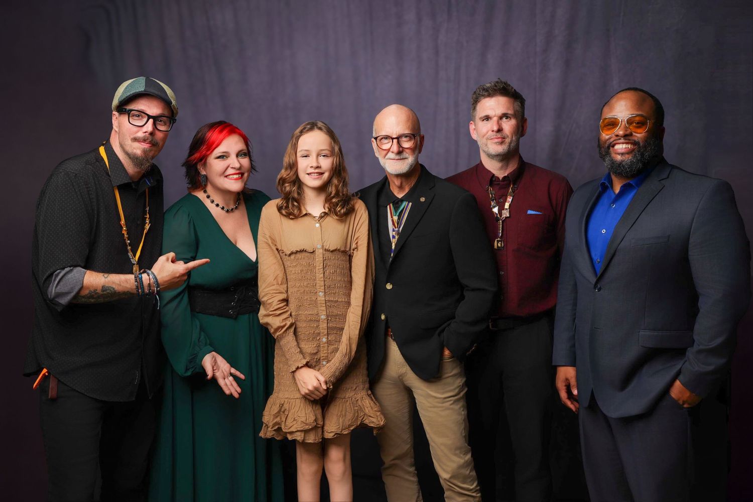 Six people posing together smiling in front of a dark gray backdrop at a professional event or premiere.