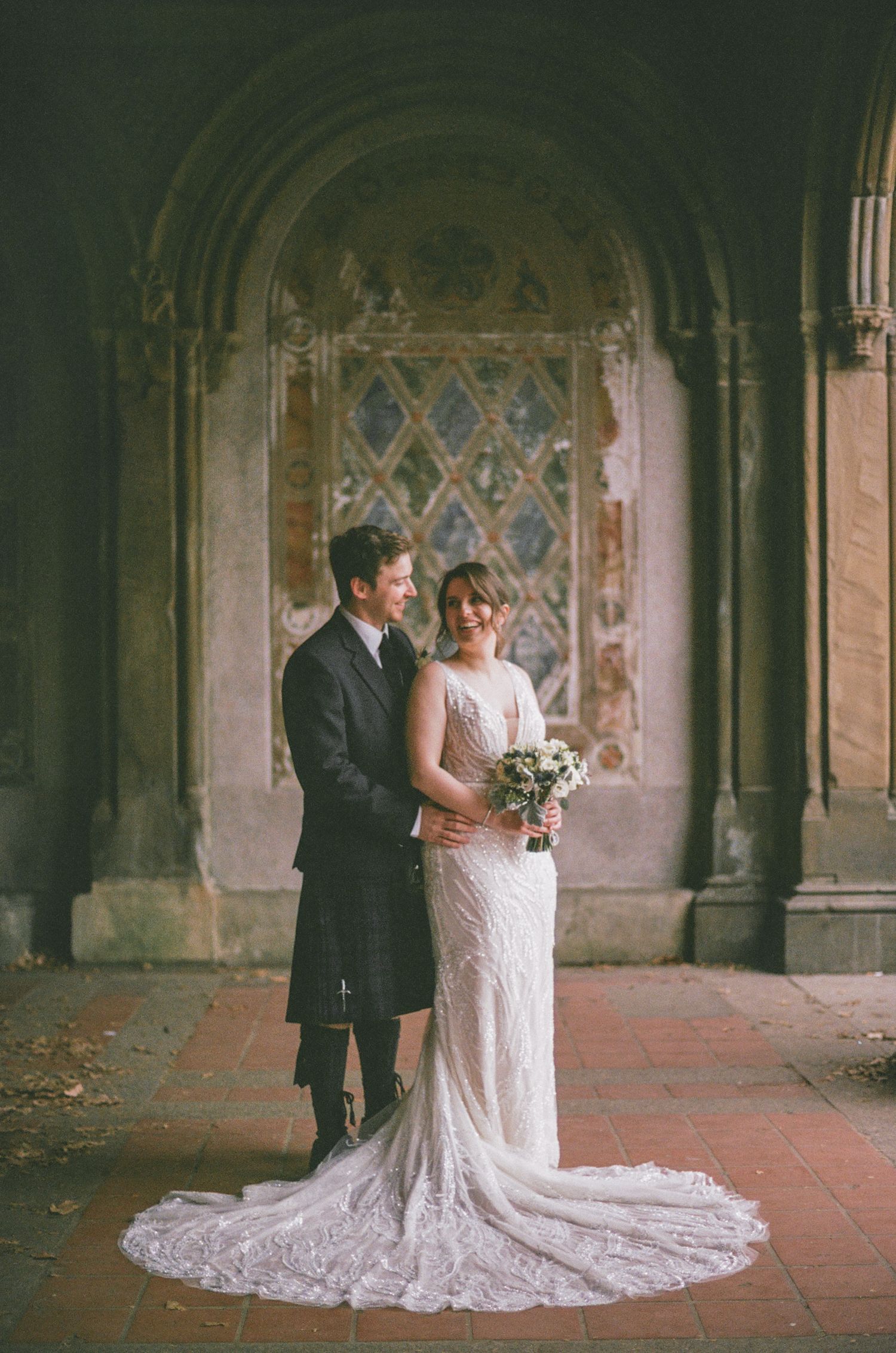 Bride in lace gown and groom in Scottish kilt pose together before ornate stained glass window at historic venue.