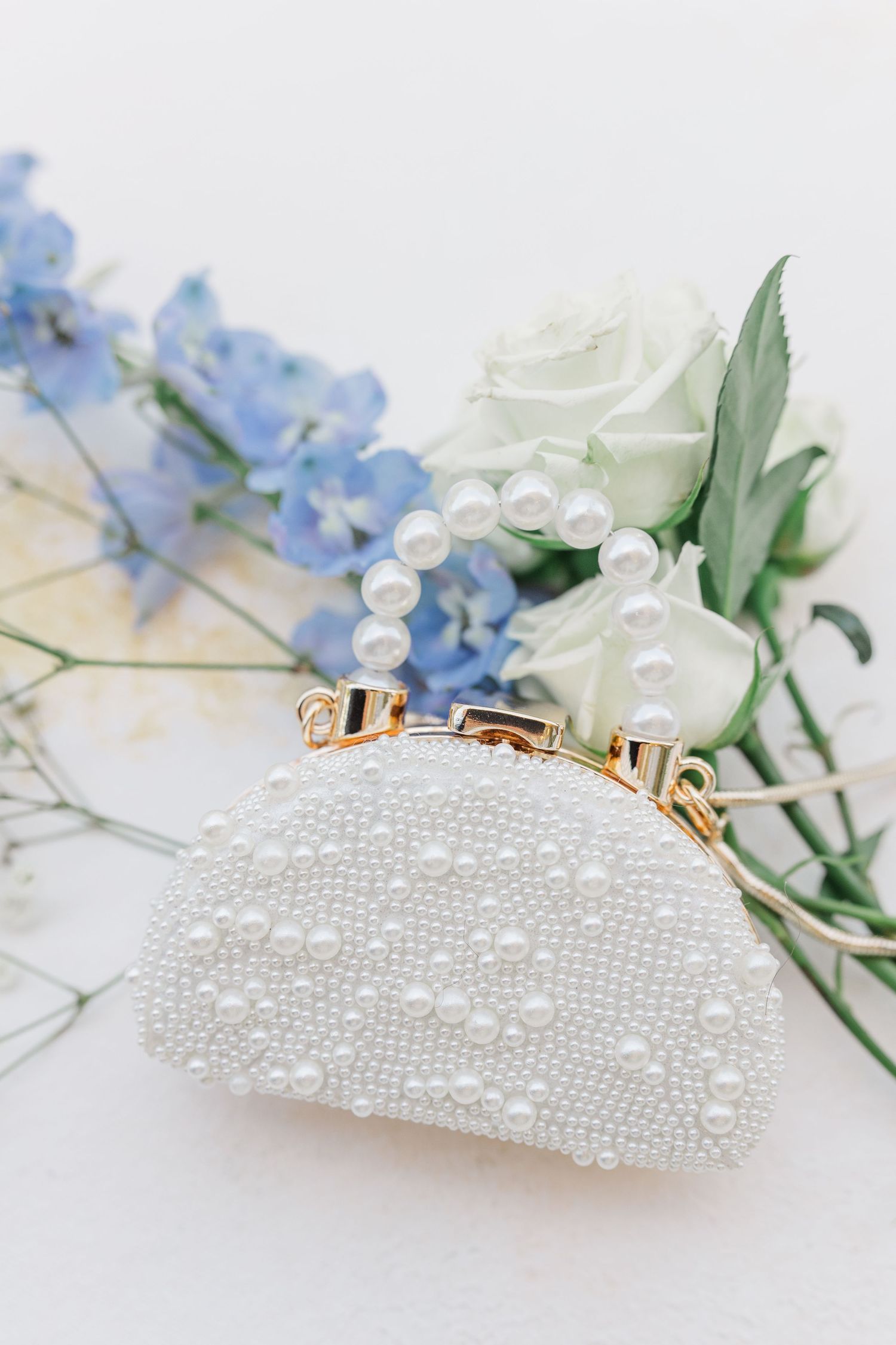 Elegant pearl-beaded white clutch purse with gold clasp beside blue hydrangeas and white rose.