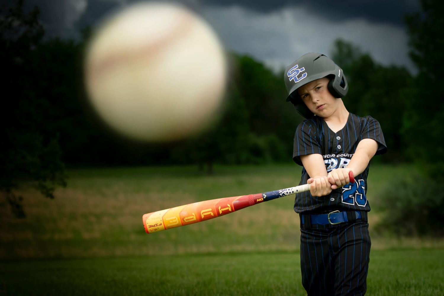 A young baseball player swings a bat at an incoming ball on a dramatic stormy field.