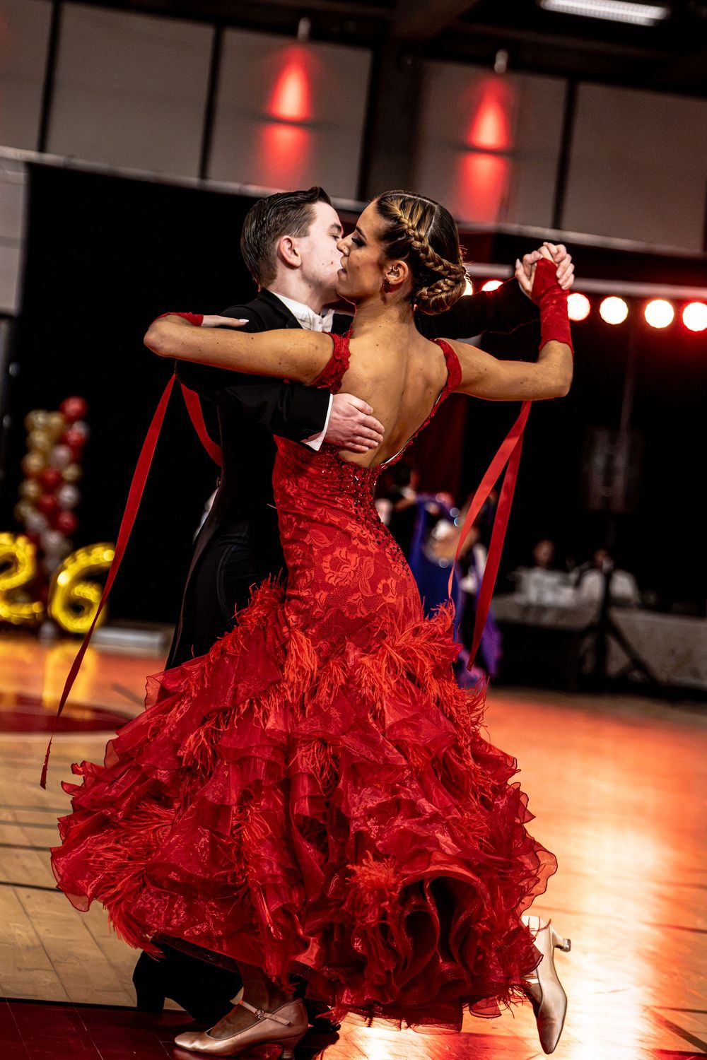 Couple performing elegant ballroom dance, woman in red ruffled gown, man in black suit, at a formal dance competition.