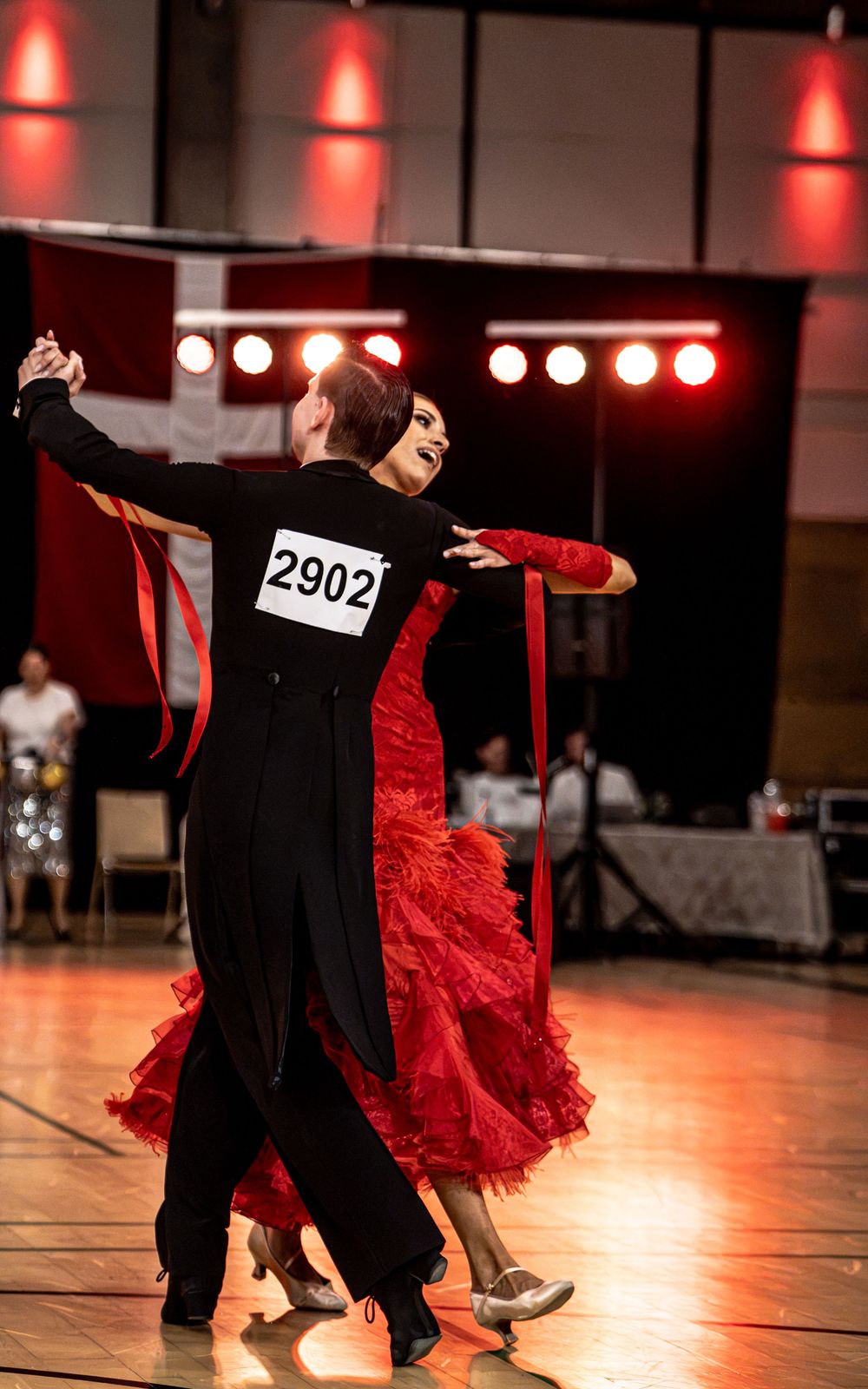 Ballroom dance couple in black and red costumes competing with number 2902 on a polished dance floor.