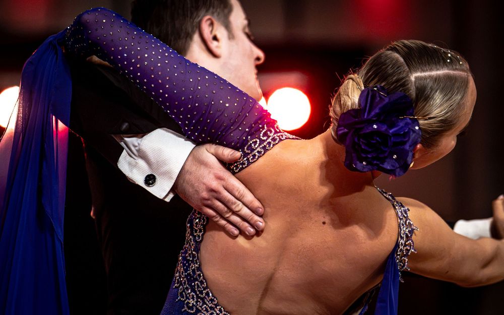 Couple ballroom dancing, woman in elegant royal blue backless gown with rhinestones and matching hair accessory under stage lights.