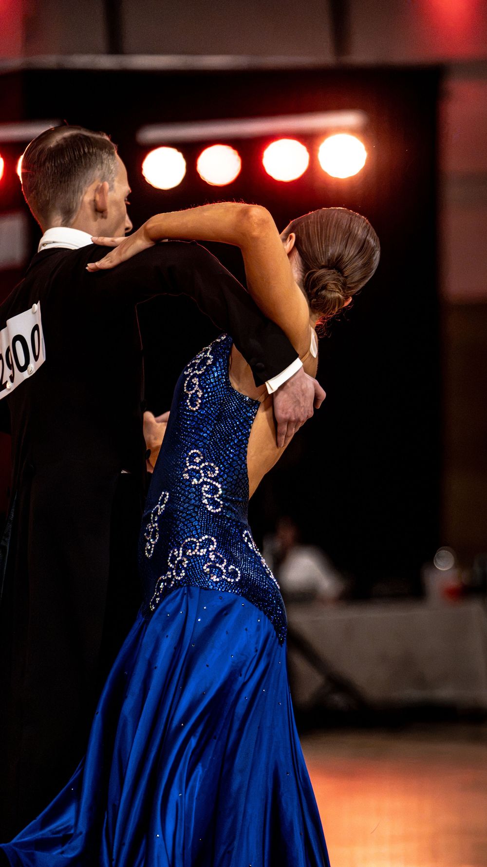Ballroom dance couple competing, woman in blue sequined gown, man in black suit, performing under stage lights.