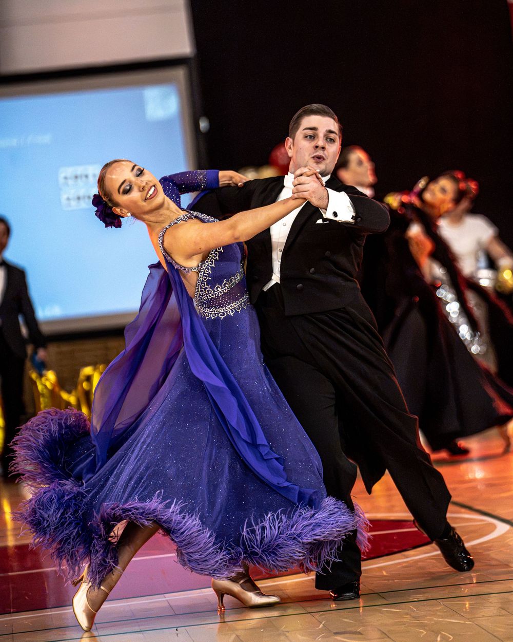 Ballroom dancers compete in elegant attire, woman in blue feathered gown and man in black tuxedo on a dance floor.