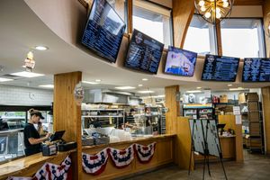Fast casual restaurant interior with digital menu boards, wood counter, patriotic bunting, and kitchen staff preparing orders.