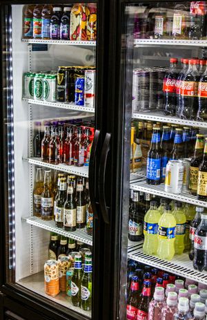 Two refrigerators stocked with assorted beverages including Coca-Cola, beer cans, and bottles in a convenience store.